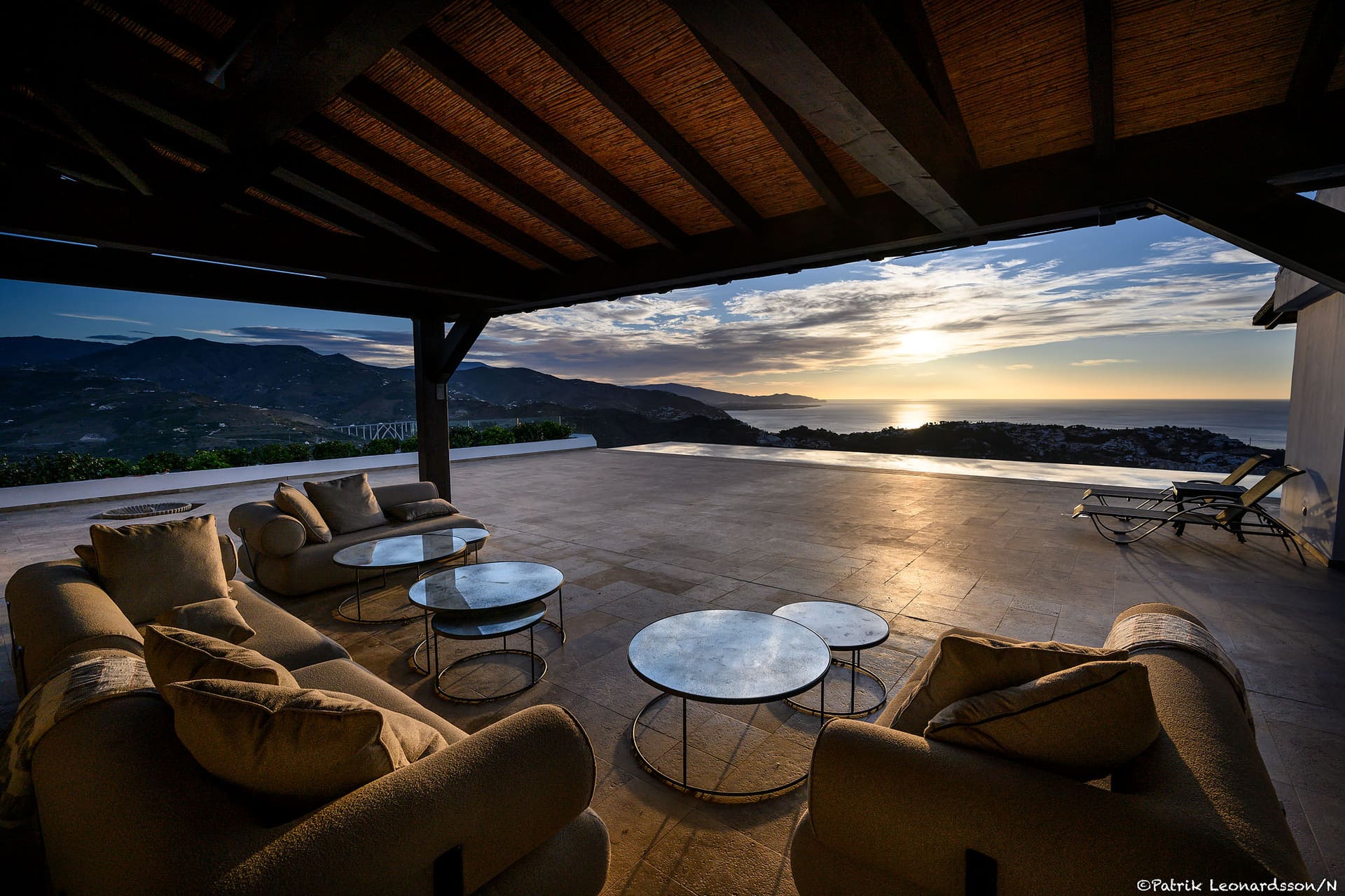 The covered terrace at sunset, sofas facing the pool's edge, the mountains and the sea.