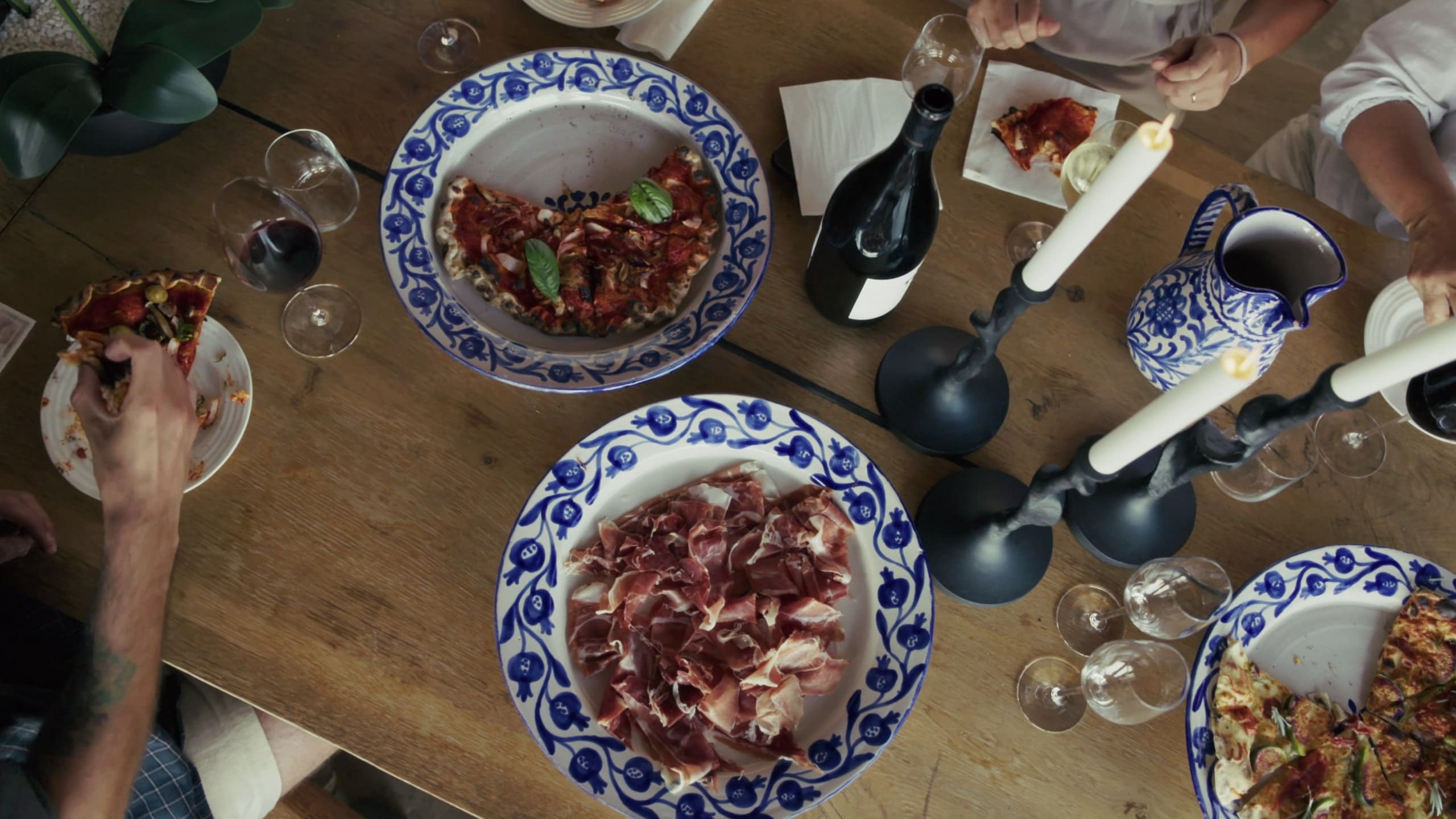 A long wooden table seen from above, plates of ham and pasta, wine and candles.