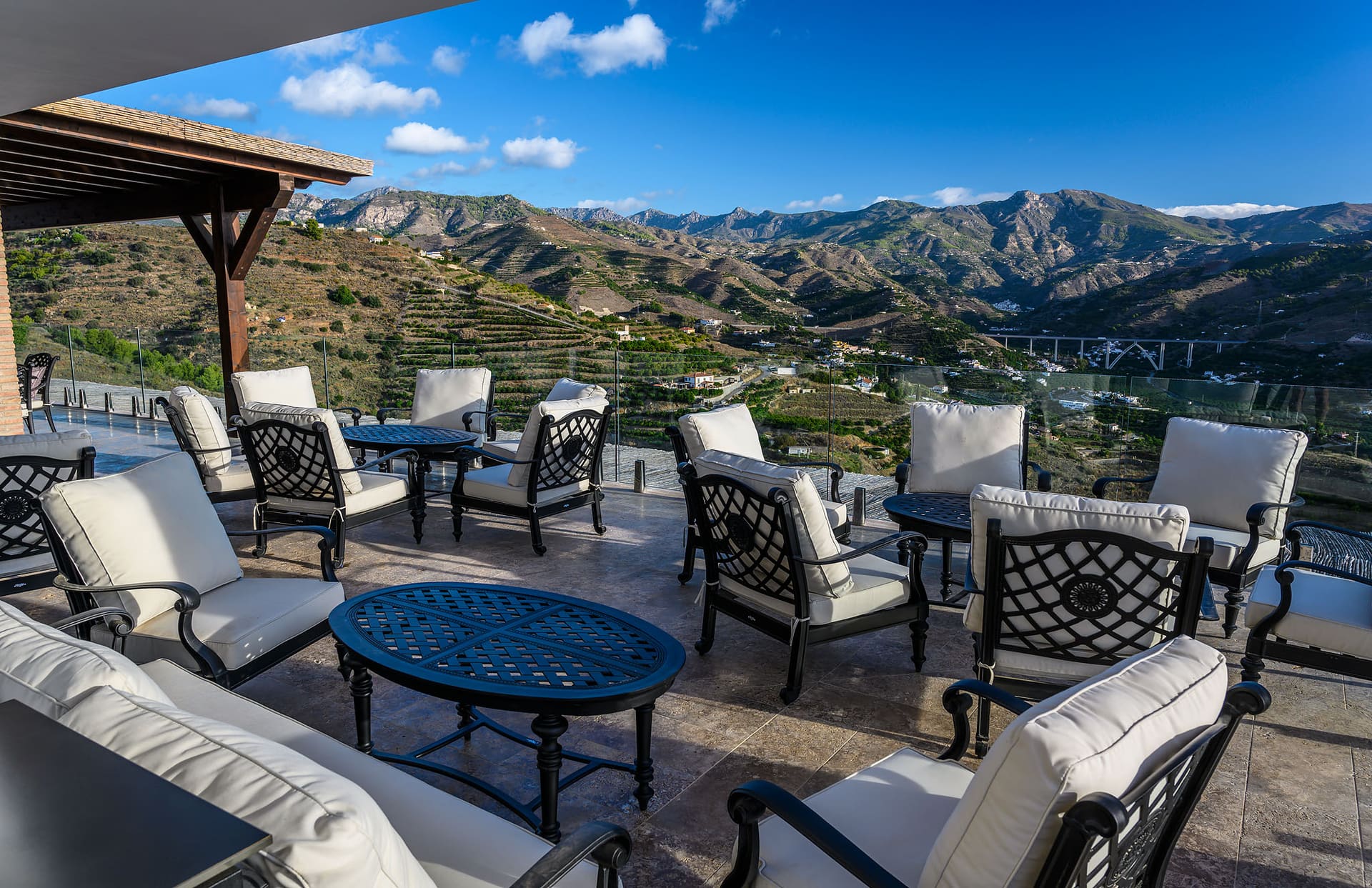 Schmiedeeiserne Stühle und Sofas auf der Terrasse unter der Pergola, dahinter die Berge.