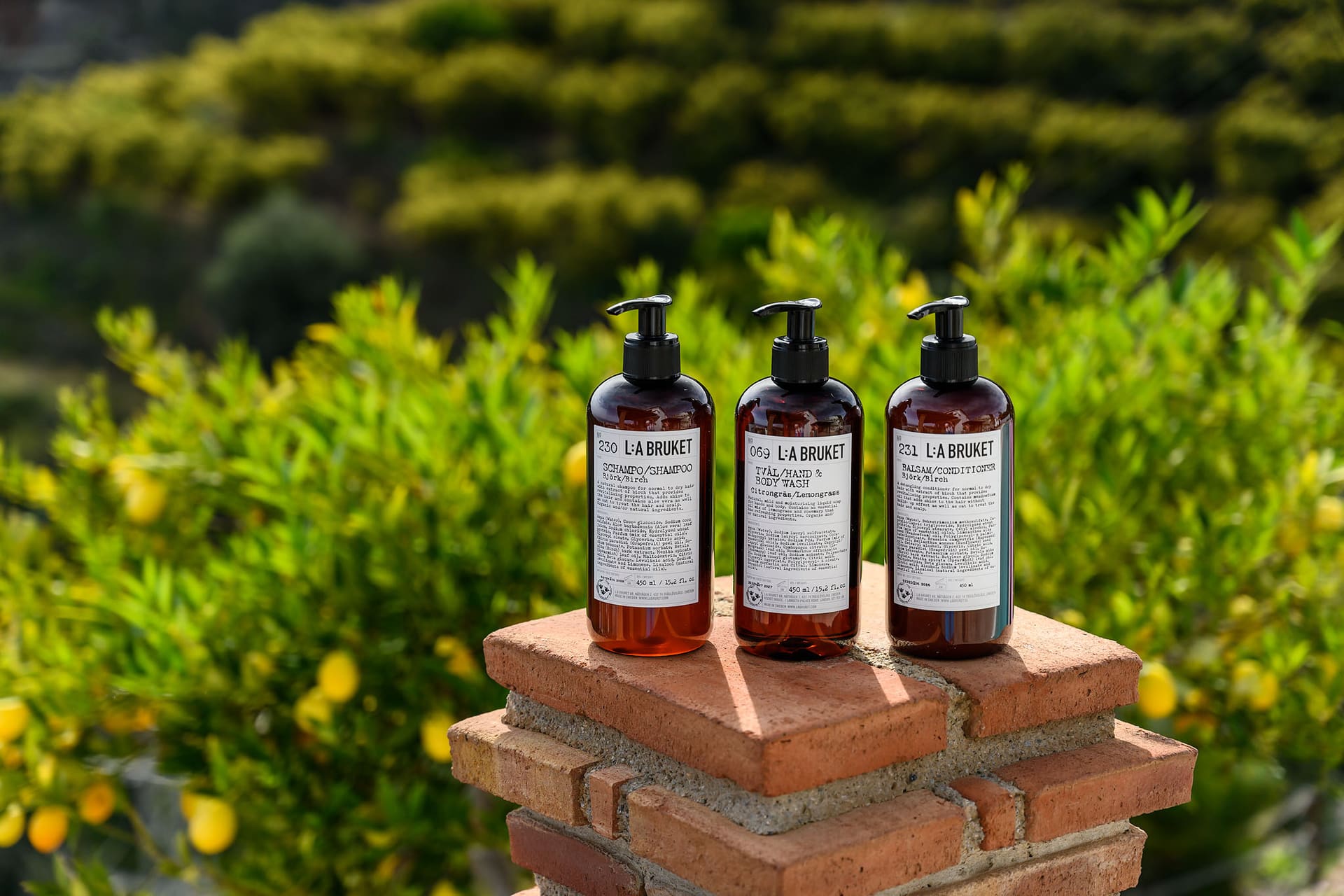 Three bottles of L:a Bruket soap on a brick pillar, lemon trees behind.