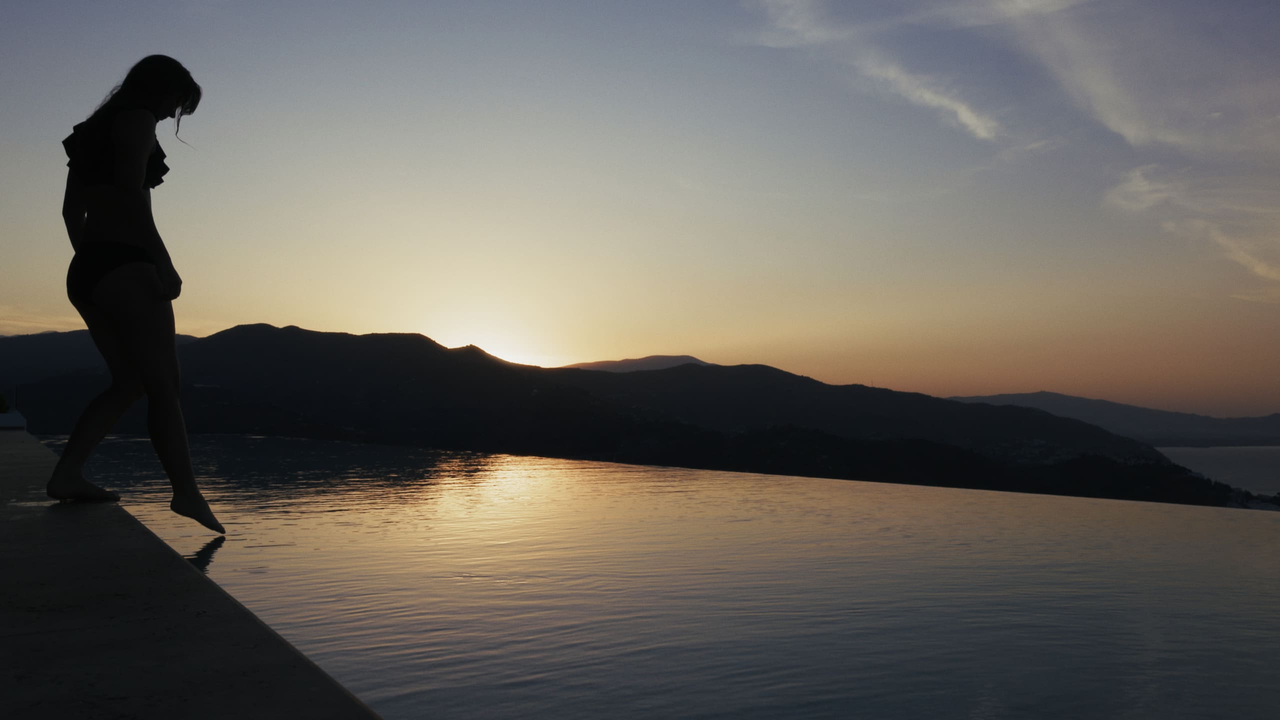 A guest at the edge of the infinity pool at sunset, the coastline silhouetted against the sky.