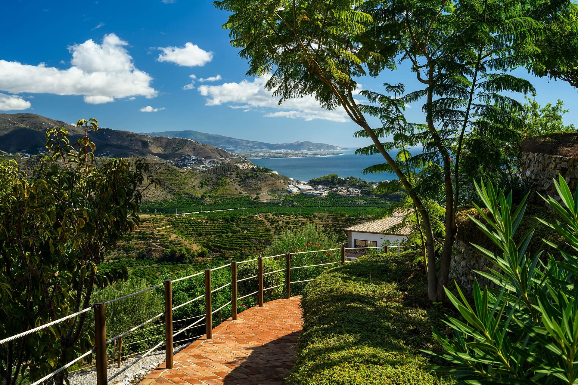A terracotta path with a wooden railing winding through the garden, terraced hills and the sea beyond.